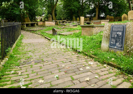 Kopfsteinpflastereingang des Friedhofs, Touristeninformationstafel, Grabsteine und Denkmäler - St. Michael & All Angels Church, Haworth, West Yorkshire, England, Großbritannien. Stockfoto