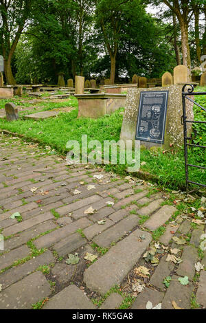 Kopfsteinpflastereingang des Friedhofs, Touristeninformationstafel, Grabsteine und Denkmäler - St. Michael & All Angels Church, Haworth, West Yorkshire, England, Großbritannien. Stockfoto