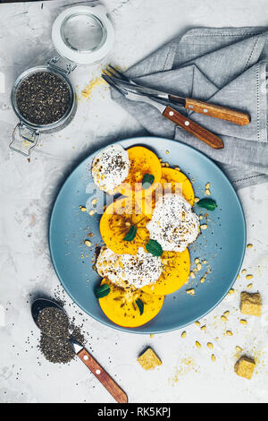 Leckere, gesunde Früchte Frühstück. Persimone Scheiben geschnitten mit Joghurt, Chia Samen, brauner Zucker, Pinienkernen und frische Minze in blauen Platte auf Hellgrau konkret Stockfoto