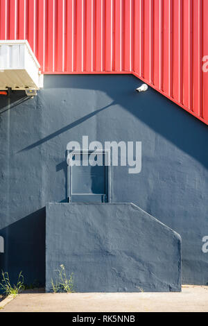 Ein fussgänger Tür in der Wand einer großen Lagerhalle, an der Spitze einer kleinen Treppe, unter einer rot Wellblech Abstellgleis mit einer Überwachungskamera. Stockfoto