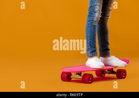 Die Beine der kleinen Mädchen in weißen Turnschuhen und Jeans auf rosa Skateboard auf orange Hintergrund Stockfoto