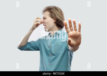 Portrait des Zurückweisens der stattlichen Langhaarigen blonden jungen Mann im blauen Hemd stehen seine Nase kneifen und jemand Blockade aufgrund schlechter Geruch. indoor St Stockfoto