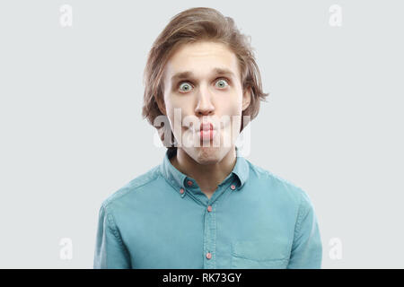 Portrait von lustig aussehenden Langhaarige, blonde junge Mann in blau casual Shirt stehen, zu Kamera mit großen Augen und Fisch Lippen suchen. indoor St Stockfoto