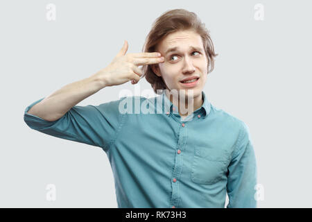 Portrait von lustig oder traurig schöne Langhaarige, blonde junge Mann in blau casual Shirt stehend mit Gewehr Pistole Geste die Hand auf den Kopf. indoor Studio s Stockfoto