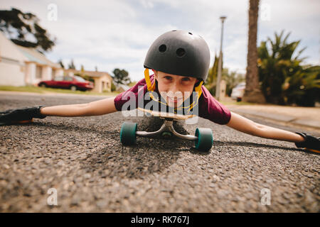 Junge mit Helm liegend auf seinem Skateboard draußen auf der Straße. Glücklich und lächelnd Junge liegend auf seinem Skateboard und Schlittschuhlaufen auf der Straße. Stockfoto