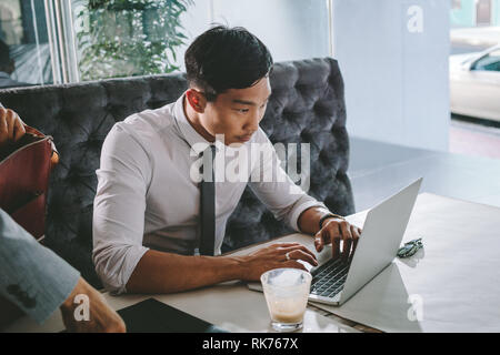 Geschäftsmann, der am Laptop arbeitet, während er im Café sitzt, und sein Kollege steht bereit. Männlicher Geschäftsmann, der an einem Laptop bei cof arbeitet Stockfoto