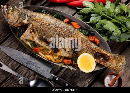 Schöner ganzer Fisch gegrillt und serviert auf einer Metallpfanne mit Tomaten, Zitrone und Kräutern Stockfoto