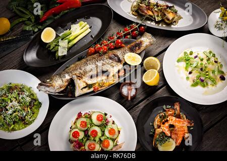 Schöner Mittagstisch mit verschiedenen Gerichten, gegrilltem Fisch, Gurkensalat, Spargel mit rasierten Parmesan und edamames Stockfoto