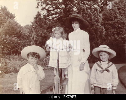 Original Vintage Cycling Edwardianer Gruppenfoto einer attraktiven Mutter in weißer Sommerbluse und Rock, die mit ihren Kindern im Park oder im Garten spazieren geht. Das jüngste Kind ist auf dem Fahrrad der Mutter ausgeglichen, die Jungs tragen modische Matrosenanzüge und Strohhüte, Edwardianerin. Edwardianerin. Die edwardianische Familie. Circa 1910, Großbritannien Stockfoto