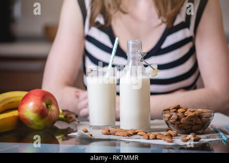 Frau trinkt organische Mandelmilch mit einem Glas in der Hand in der Küche. Flasche und Obst auf den Tisch. Gesunde vegetarische Produkt Stockfoto