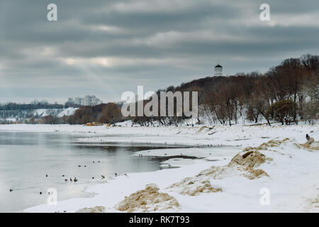 Blick auf den zugefrorenen Fluss mit Raben auf dem Eis und das Stadtbild im Hintergrund, während Sie im malerischen Gesägt an einem Wintertag in Kolomenskoye Park, Moskau Stockfoto