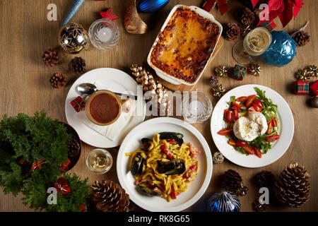 Flaches Foto von einem Weihnachtstisch mit Lasagne, Muschelpasta, Burrata-Salat und einem Tiramisu, serviert mit etwas Sekt Stockfoto