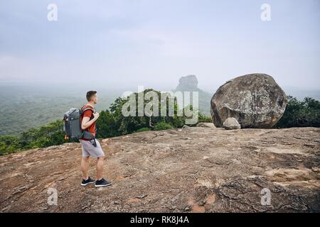 Junger Mann mit Rucksack gegen Sigiriya Felsen (UNESCO-Weltkulturerbe). Blick von pidurangala Felsen in Sri Lanka. Stockfoto