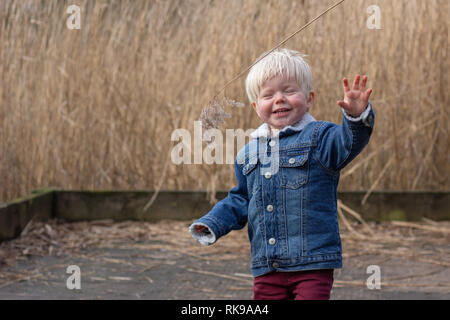 Eine blonde junge männliche Kleinkind genießen der Park und das Lächeln, die versucht, für langes Gras zu erreichen. Stockfoto