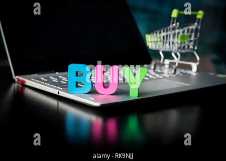 Kreative Schuß eines Holz- viele - farbige Wort kaufen auf Laptop unten an der schwarzen Bildschirm und Spielzeug Einkaufswagen Hintergrund. Konzept der kaufen Stockfoto