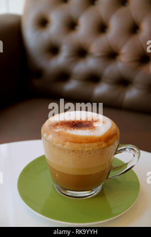 Tasse fluffigen Milchschaum heißen Cappuccino am weißen Tisch mit verschwommenen Sofa im Hintergrund Stockfoto