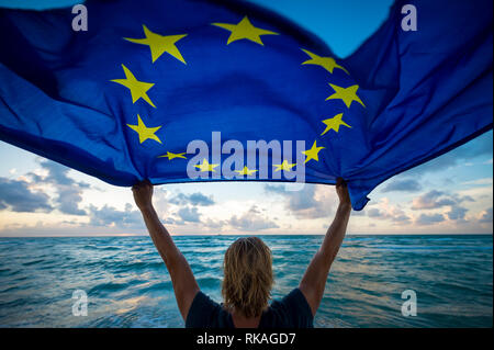 Mann mit einer flatternden Fahne der Europäischen Union in den hellen Morgen Sonnenlicht durch Mann mit blonden Haaren auf leeren Strand am Mittelmeer statt Fliegen Stockfoto