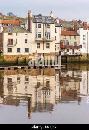 Gebäude entlang der Uferpromenade am Whitby im Wasser des Hafens wider. Ein Hügel ist hinter den Häusern und einem blauen Himmel. Stockfoto