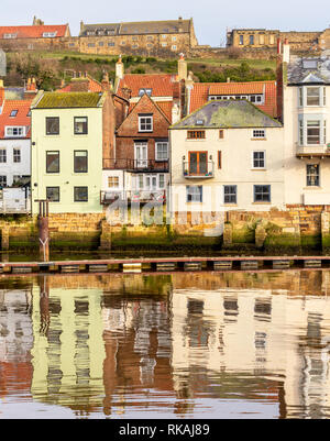 Gebäude entlang der Uferpromenade am Whitby im Wasser des Hafens wider. Ein Hügel ist hinter den Häusern und einem blauen Himmel. Stockfoto