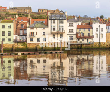 Gebäude entlang der Uferpromenade am Whitby im Wasser des Hafens wider. Ein Hügel ist hinter den Häusern und einem blauen Himmel. Stockfoto