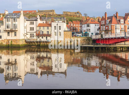 Gebäude entlang der Uferpromenade am Whitby im Wasser des Hafens wider. Ein Hügel ist hinter den Häusern und einem blauen Himmel. Stockfoto