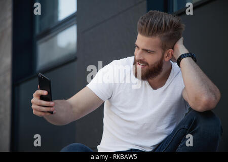 Gut aussehender bärtiger junger Mann macht selfie und lächelt er im weißen T-Shirt gekleidet ist Stockfoto