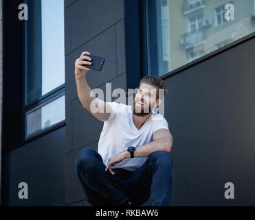 Gut aussehender bärtiger junger Mann macht selfie und lächelt er im weißen T-Shirt gekleidet ist Stockfoto