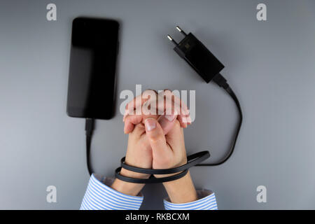 Weibliche Hände und Smartphone. Woman's Händen gefangen und der Handgelenke mit Handy Kabel wie Handschellen gewickelt. Neigung zu Internet und soziale Netzwer Stockfoto