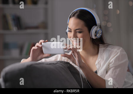 Glückliche Frau Uhren videos mit Kopfhörern sitzen auf einer Couch in der Nacht zu Hause Stockfoto