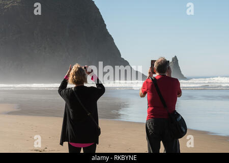 Ein paar Touristen nehmen Bilder mit ihren Telefonen und Tabellen Haystack Rock in Cannon Beach, Oregon, USA. Stockfoto
