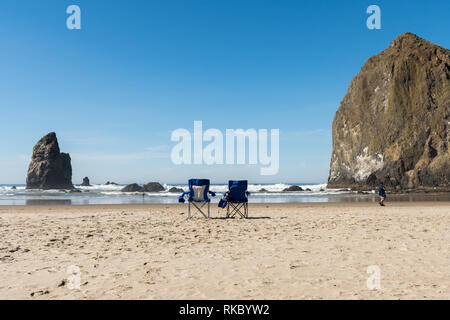 Zwei blaue Klappstühle in den Sand als touristische Spaziergänge entlang des Strandes von Cannon Beach, Oregon, USA. Stockfoto