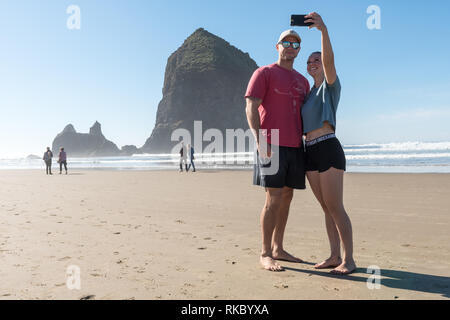 Ein junges Paar von Touristen machen selfies vor dem Haystack Rock in Cannon Beach, Oregon, USA. Stockfoto