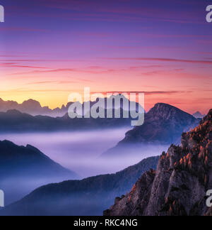 Berge im Nebel bei Sonnenuntergang im Herbst in den Dolomiten, Italien. Landschaft mit Alpine Mountain Valley, niedrige Wolken, Bäume auf Hügeln, lila Himmel mit Stockfoto