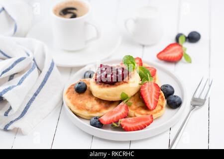 Quark Pfannkuchen oder Syrniki mit Erdbeeren, Heidelbeeren, Beeren Marmelade und Tasse schwarzen Kaffee Espresso. Leckeres Frühstück am weißen Tisch Stockfoto