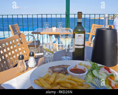 Schwertfisch mit Pommes und Salat und eine Flasche Weißwein der Maltese Falcon, den Blick auf das Mittelmeer in Bugibba St. Paul's Bay, Malta Stockfoto