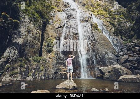 Junger Mann (touristische) stehen auf Stein in und Strom des fallenden Wassers mitten in der Natur pur. Bambarakanda Wasserfall - der höchste Wasserfall Stockfoto