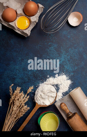 Zutaten und Utensilien zum Backen. Löffel mit Mehl, Eiern, Nudelholz, Backpapier, Schneebesen für Peitschen auf blauem Grund. Ansicht von oben, selektive f Stockfoto