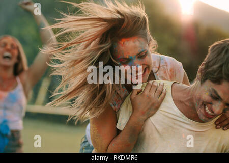 Lächelnde Frau Spaß haben mit ihrem Freund beim Spielen Holi in einem Park. Frau mit ihrem Mann von hinten während des Spielens Holi mit Freunden zujubeln i Stockfoto