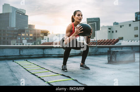 Weibliche Athleten dabei hockt Halte einen Medizinball auf einem Dach. Frau tun Trainings mit Medizinball mit einer Agilität Leiter durch ihre Seite Stockfoto