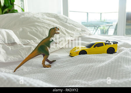 Spielzeug Dinosaurier und Auto auf dem Bett der Eltern, Familie und Elternschaft leben. Weiße Bettwäsche im Hintergrund. Stockfoto