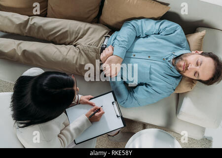 Ansicht von oben Der psychotherapeut Schreiben auf die Zwischenablage und männlichen Patienten mit geschlossenen Augen auf der Couch liegend Stockfoto