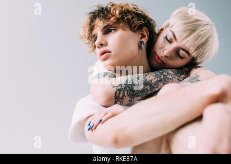 Blonde Frau mit Tattoos umarmen Mann mit lockigem Haar isoliert auf Grau Stockfoto