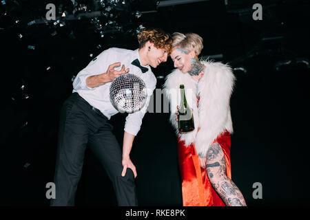 Mann hält Disco ball und stehen in der Nähe von Frau mit Flasche Alkohol trinken auf Party auf schwarzem Hintergrund Stockfoto