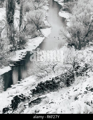 Eine verschneite Landschaft, halb zugefrorenen Fluss mit Bäumen an den Ufern, fließt durch ein flaches Tal. Die Schwarz-Weiß-Fotografie Stockfoto