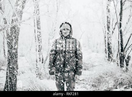 Ein Kerl, der eine Jacke mit Kapuze steht in einem verschneiten Wald, Schnee fällt auf ihn von oben. Schwarz und Weiß Foto Stockfoto