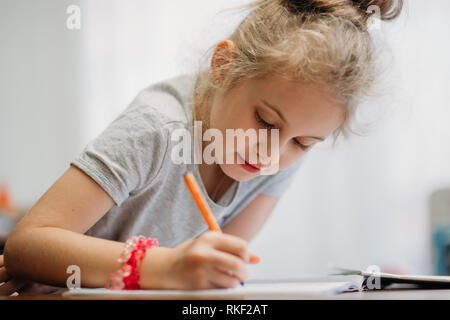 Ein siebenjähriges Mädchen sitzt zu Hause am Tisch und schreibt in einem Notebook, Abschluss einer Aufgabe lernen oder wiederholen. Stockfoto
