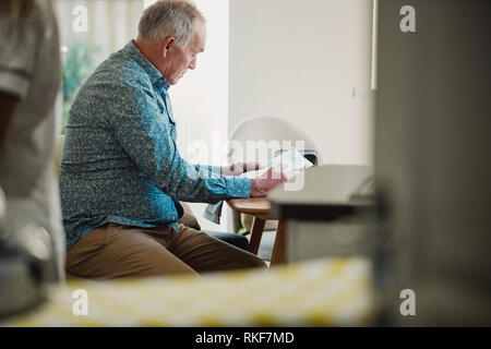 Ältere Diabetiker Mann sitzt an einem Esstisch eine Zeitung lesen, während seine Pflegeperson seine Medikamente bereitet. Stockfoto
