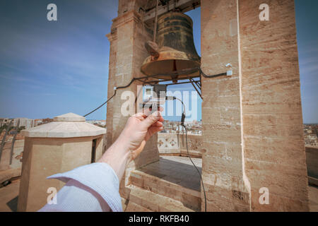 Ein Mann hält eine GoPro Kamera vor einer großen Glocke an der Spitze der El Miguelete, der Glockenturm der Kathedrale von Valencia in Valencia, Spanien. Stockfoto