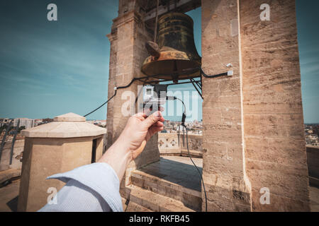 Ein Mann hält eine GoPro Kamera vor einer großen Glocke an der Spitze der El Miguelete, der Glockenturm der Kathedrale von Valencia in Valencia, Spanien. Stockfoto