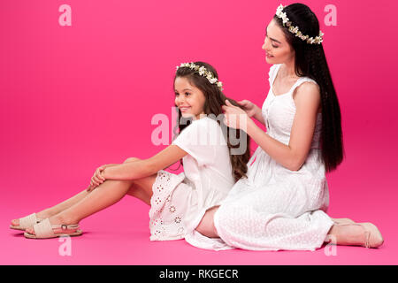 Glückliche junge Mutter flechten Braid zu schönen lächelnden Tochter auf Rosa Stockfoto